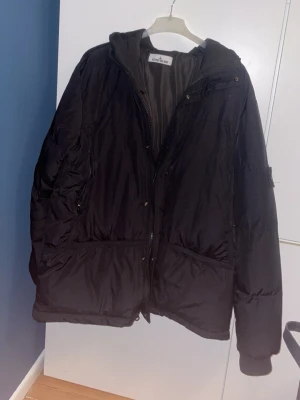 Svart pufferjacka från Stone Island - Säljer en svart pufferjacka från Stone Island med ikonisk patch på ärmen. Jackan har huva, dragkedja framtill och stora fickor. Tillverkad i nylon med Tyvek-sköld som gör den vind- och vattenavvisande. Perfekt för kalla dagar och riktigt snygg streetstil.