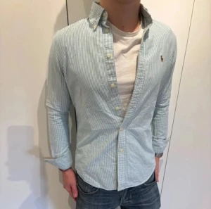 Randig skjorta från Polo Ralph Lauren - Klassisk blå- och vitrandig skjorta från Polo Ralph Lauren i bomull. Skjortan har lång ärm, knappar framtill och den ikoniska broderade loggan på bröstet. Classic fit med stilren krage, perfekt för en clean och tidlös look.
