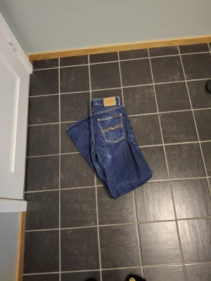 Blå jeans från Nudie Jeans, W30 L32 - Säljer ett par klassiska blå jeans från Nudie Jeans med raka ben och kontrastsömmar. Jeansen har fem fickor och en snygg detalj på bakfickan. Tillverkade i slitstarkt bomullsmaterial och passar dig som gillar tidlös denimstil.