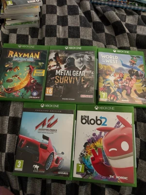 Xbox One Spelpaket (5 spel) - Fem Xbox One-spel: Rayman Legends, Metal Gear Survive, World to the West, Assetto Corsa och de Blob 2. Skivorna och fodralen ser ut att vara i gott skick, perfekta för samlare eller spelentusiaster. Inga synliga defekter.