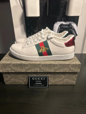 Gucci Ace sneakers med bi-detalj - Säljer ett par Gucci Ace sneakers i vitt skinn med klassiska gröna och röda ränder på sidan och broderad guldfärgad bi. Baksidan har metallicröd och metallicgrön häl med ormmönster och Gucci-logga. Platt sula med Gucci-mönster undertill och snörning framtill.