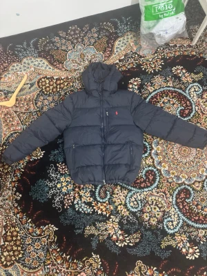 Mörkblå pufferjacka från Polo Ralph Lauren - Säljer en mörkblå pufferjacka från Polo Ralph Lauren med huva och dragkedja framtill. Jackan har två sidofickor med dragkedja och den klassiska röda Polo-loggan på bröstet. Perfekt för kalla dagar och riktigt snygg till streetwear-stilen.