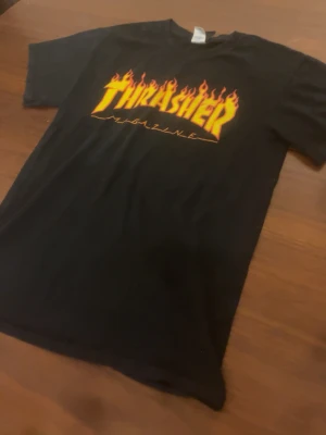 Svart Thrasher Flame t-shirt - Klassisk svart Thrasher t-shirt med den ikoniska Flame-loggan i gult och rött på bröstet. Skön passform och tillverkad i mjuk bomull. Perfekt för dig som gillar streetwear och skatekultur. Den är storlek S men skulle säga passar lite mer som en xs