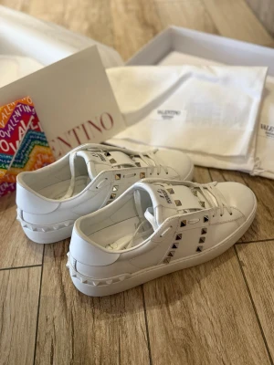 Valentino Rockstud vita sneakers - Säljer ett par vita Valentino Rockstud sneakers i skinn med ikoniska nitar i silver och guld längs sidorna och på tungan. Klassisk rund tå, snörning och platt sula med mönstrad undersida. Kommer med originalkartong, dustbag och extra skosnören.