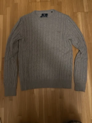 Grå kabelstickad tröja från Gant,  - Kan går ner ordentligt i pris vid snabb affär. Säljer en klassisk grå kabelstickad tröja från Gant med rund hals och diskret logga på bröstet. Tröjan har långa ärmar och ribbade muddar vid ärmslut och nederkant. Perfekt för lager på lager under hösten och vintern.