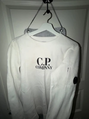 Vit sweatshirt från C.P. Company Äkta Storlek S/M - Vit sweatshirt från C.P. Company med svart logga på bröstet och ikonisk linsdetalj på ärmen. Tröjan har lång ärm, rund hals och en ficka med dragkedja på ärmen. Tillverkad i mjuk bomull för skön känsla.