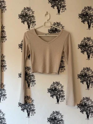 Beige ribbad croppad långärmad topp - Trendig beige croppad topp med lång ärm och v-ringning. Tillverkad i ribbad bomullsmix som ger en snygg och bekväm passform. Perfekt att styla med höga jeans eller kjol för en clean look.