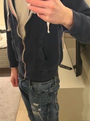 Mörkblå hoodie med dragkedja - Mörkblå hoodie från Style Level med vit dragkedja och vita snören i huvan. Mjuk insida och två fickor framtill. Perfekt för en avslappnad look och enkel att matcha med jeans eller joggers.