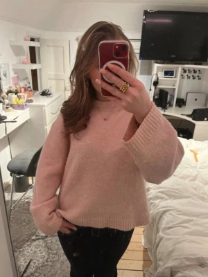 Ljusrosa stickad tröja - Mysig ljusrosa stickad tröja med breda ärmar och ribbade muddar. Tröjan har en avslappnad passform och är perfekt för kalla dagar. Materialet ser ut att vara mjukt och fluffigt, vilket ger en skön känsla.
