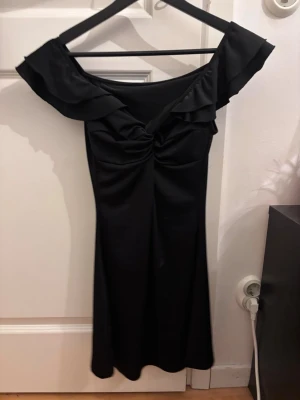 Svart offshoulder klänning med volang - Stilren svart klänning med offshoulder och volangdetaljer upptill. Klänningen har en figurnära passform och är tillverkad i ett mjukt, stretchigt material. Perfekt för dig som vill ha en elegant look med en twist.