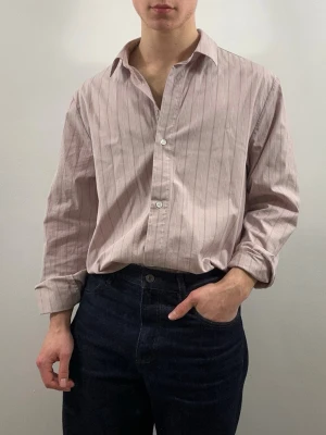 COS Skjorta Ljusrosa Randig M - Snygg ljusrosarandig skjorta från COS med tunna vertikala ränder och klassisk krage. Modellen är 175 cm och väger 70 kg. M i regular fit. Skjortan har lång ärm, knappar framtill och en avslappnad passform. Perfekt för en stilren och trendig look.
