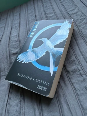 Revolt av Suzanne Collins - Säljer boken 'Revolt' av Suzanne Collins, tredje delen i Hungerspelen-serien. Omslaget är svart med en blå fågel och text i blått och vitt. Pocketbok med mjuka pärmar, tryckt på papper. Perfekt för dig som gillar dystopisk science fiction.