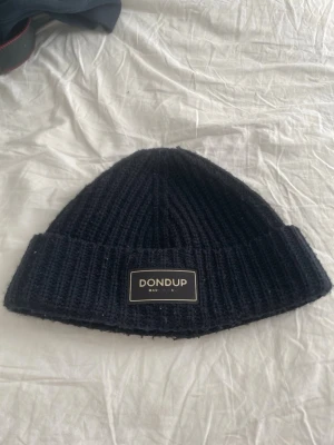 Svart ribbstickad mössa från Dondup - Säljer en svart ribbstickad mössa från Dondup med uppvikt kant och en snygg logotyp-patch framtill. Klassisk modell som passar perfekt till höst och vinter. Materialet känns mjukt och värmande, perfekt för kalla dagar. Texten under dondup har gått bort lite det ska stå made in Italy. Köpt för 1000 kr säljer för 350 