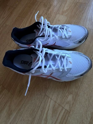 Vita Asics sneakers med mesh och snörning - Snygga vita Asics sneakers med mesh-ovandel och detaljer i silver och rött. Skorna har klassisk snörning och svart innersida. Perfekta för träning eller en sportig look. Lätta och ventilerande material gör dem bekväma att använda hela dagen.