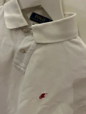 Vit pikétröja från Polo Ralph Lauren - Klassisk vit pikétröja från Polo Ralph Lauren med korta ärmar och krage. Tröjan har knäppning framtill och den ikoniska röda logotypen broderad på bröstet. Perfekt till jeans eller shorts för en clean look. Fint skick förutom att den är något skrynklig på bilderna men det är lätt att fixa.