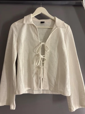 Vit topp med snörning från Gina Tricot - Säljer en vit långärmad topp från Gina Tricot med snygg snörning framtill och krage. Toppen har en lätt struktur i tyget och är croppad i modellen. Perfekt att styla med jeans eller kjol för en trendig look.