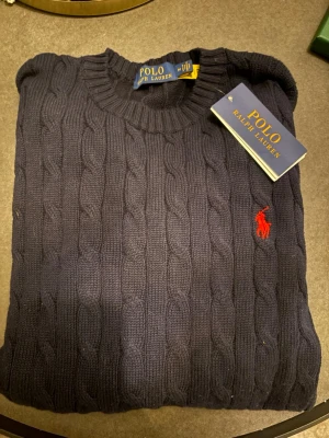 Mörkblå kabelstickad tröja Polo Ralph Lauren - Kabelstickad mörkblå tröja från Polo Ralph Lauren med rund halsringning och klassisk röd broderad logga på bröstet. Tröjan är i mjuk stickad bomull och har ribbade muddar vid ärmslut och nederkant. Perfekt för en stilren look. Osäker på äkthet då jag fick den som en present