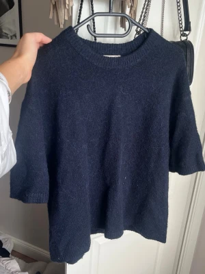 Mörkblå stickad t-shirt - Mysig mörkblå stickad t-shirt med korta ärmar och rund halsringning. Tröjan har en avslappnad passform och ribbstickade detaljer vid hals och ärmslut. Perfekt för lager-på-lager och enkel att styla till olika outfits.