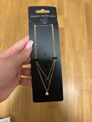 Guldigt dubbelt halsband Design Jewellery - Snyggt dubbelt halsband i guldfärgad metall från Design Jewellery. Ena kedjan har en liten cirkelberlock och den andra en klar stenliknande detalj. Perfekt lager-på-lager-look med tunna kedjor och minimalistisk design.