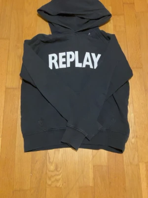 Svart hoodie från Replay - Svart hoodie från Replay med stor vit logga på bröstet. Klassisk modell med huva och ribbade muddar vid ärmslut och nederkant. Perfekt för en avslappnad och cool stil. Materialet känns mjukt och bekvämt mot huden.