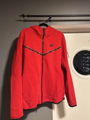 Röd Nike tech hoodie - Snygg röd hoodie från Nike med dragkedja. Finns en skada på framsidan och en fläck vid ena armen men annars bra skick