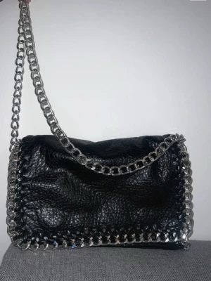 TIAMO svart väska - Svart väska med silverfärgade kedjedetaljer. Väskan har ett innerfack med dragkedja och en liten ficka.  Jätte fin, välskött crossbody väska utan skador. Det är i den större storleken