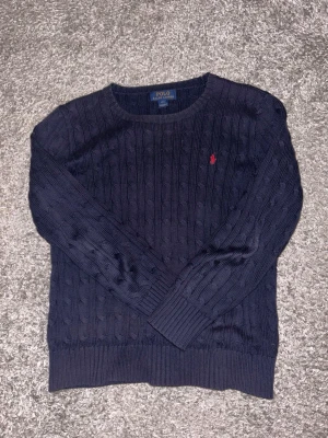 Raulph lauren stickad tröja - Kabelstickad mörkblå tröja från Polo Ralph Lauren med rund halsringning och röd broderad logga på bröstet. Tröjan har ribbade muddar vid ärmslut och nederkant. Perfekt för lager-på-lager och klassisk stil. Passar 160-170 cm lång person