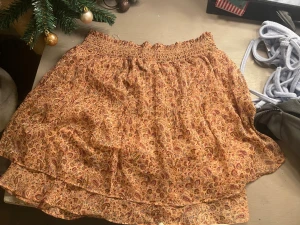 Blommig kortkjol från Neo Noir - Supersöt kortkjol från Neo Noir i ett blommigt mönster med varma toner av beige, röd och orange. Kjolen har smock i midjan och dubbla lager för en flowy känsla. Perfekt för dig som gillar en feminin och avslappnad stil.