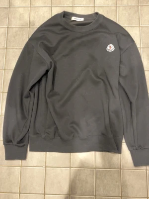 Svart sweatshirt från Moncler - Säljer en svart sweatshirt från Moncler med rund hals och ribbade muddar. Tröjan har Moncler-logga på bröstet och ett stort vitt M-tryck på ryggen. Perfekt för dig som gillar stilrena och exklusiva plagg. Passar till storlek S och M