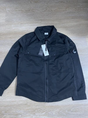 Svart overshirt från CP - Stilren svart overshirt  CP med dragkedja framtill, två bröstfickor med knappar och klassisk krage. Ärmen har den ikoniska linsen på sidan. Perfekt lager för en modern look.