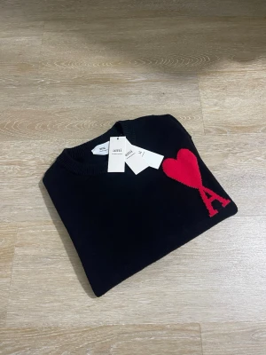 Svart stickad tröja  - Svart stickad tröja med rund halsringning och ett rött hjärta med bokstaven A broderat på bröstet. Tröjan har ribbstickade muddar och är i en klassisk, stilren design.