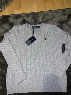 Grå kabelstickad tröja Polo Ralph Lauren - Snygg grå kabelstickad tröja från Polo Ralph Lauren med rund halsringning och klassisk broderad logga på bröstet. Tröjan har långa ärmar och ribbade muddar vid ärmslut och nederkant. Perfekt för lager-på-lager och stilren look.storlek S ,slim !  kan vara lite tight om du är över 170! Helt ny aldrig använd , 