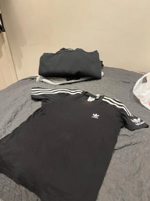 Svart Adidas t-shirt med vita ränder - Snygg svart t-shirt från Adidas med klassiska vita ränder på axlarna och ärmarna. Adidas logga tryckt på bröstet och ärmen soppar till en sportig vibe. Perfekt till streetwear och casual outfits. Tillverkad i mjukt bomullsmaterial för skön känsla.