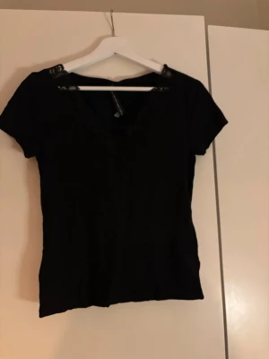 Svart topp med spets  - Snygg svart topp från med v-ringning och spetsdetaljer vid halsen. Kortärmad och figurnära modell som passar perfekt till jeans eller kjol. Enkel men ändå elegant design.