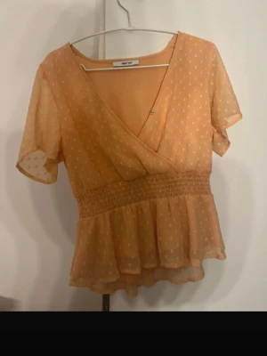 Prickig omlottopp från About You - Söt persikofärgad omlottopp från About You med korta ärmar och prickigt mönster. Toppen har smock i midjan och volang nedtill för en feminin look. Perfekt till jeans eller kjol för en trendig outfit.