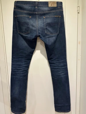 Nudie jeans Grim Tim - Säljer ett par Nudie jeans Grim Tim i mörkblå tvätt. Modellen har en slim passform med raka ben och fem fickor. Snygga kontrastsömmar och diskret läderpatch bak i midjan. Perfekta till vardagslooken!