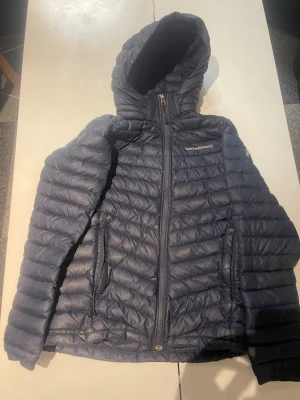 Blå dunjacka från Peak Performance - Snygg blå dunjacka från Peak Performance med huva och dragkedja framtill. Jackan har quiltad design, två sidofickor med dragkedja och logga på bröstet. Perfekt för kalla dagar och enkel att matcha med olika outfits.