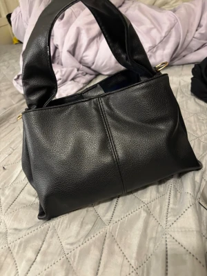 Svart handväska i skinnimitation - Stilren svart handväska med bred axelrem och guldfärgade detaljer. Väskan är tillverkad i skinnimitation och har ett rymligt fack med röd textilinsida och magnetknäppning. Perfekt för dig som vill ha en enkel men snygg accessoar till vardagslooken.