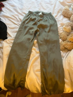 Olivgrön satinpyjamas från H&M - Säljer ett set med olivgröna pyjamasbyxor och matchande skjorta från H&M. Båda delarna är i mjukt, glansigt satintyg och har en avslappnad passform. Skjortan har knappar framtill och klassisk krage, byxorna har resår i midjan för extra komfort.