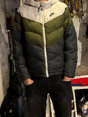 Nike pufferjacka i vitt, grönt och svart - Nike pufferjacka med färgblock i vitt, olivgrönt och svart. Jackan har huva, dragkedja framtill och klassisk quiltad design. Perfekt för kalla dagar och ger en sportig vibe. Nike-logga på bröstet och normal passform.