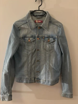 Ljusblå klassisk jeansjacka - Snygg ljusblå jeansjacka i klassisk modell med krage och knappdetaljer. Tillverkad av 98% bomull och 2% elastan för en bekväm passform. Perfekt till vardagslooken och enkel att matcha med olika outfits.