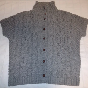 Grå/beige/brun stickad väst/kortärmad tröja/ kofta med knappar - Mysig grå/beige/brun stickad kofta från Hampton Republic med korta ärmar och hög ribbad krage. Koftan har ett snyggt flätmönster och bruna knappar framtill. Perfekt för lager-på-lager och chill höst- och vinterstil. Storlek 36/38. Är lite nopprig.