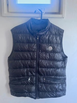  Moncler väst - Snygg svart dunväst från Moncler med hög krage och dragkedja framtill. Västen har två framfickor med dragkedja och det klassiska Moncler-logotypen på bröstet. Tillverkad i glansigt material med quiltad design som ger en modern look.