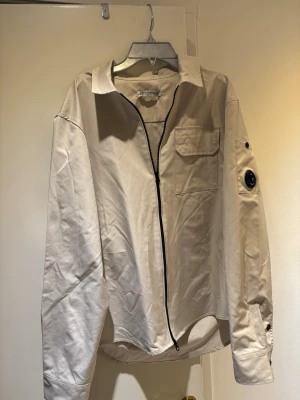 Beige overshirt jacka från C.P. Company - Stilren beige overshirt jacka från C.P. Company med svart dragkedja framtill, bröstficka och ikonisk linsdetalj på ärmen. Tillverkad i bomull med klassisk krage och långa ärmar. Perfekt för en avslappnad men snygg look.