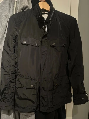 Svart vindjacka med fickor - Snygg svart vindjacka med fyra framfickor med lock och knappar, hög krage och dragkedja framtill. Jackan har en klassisk passform och är tillverkad i ett lätt, vindtätt material som passar perfekt för blåsiga dagar. Sparsamt använd, storlek M