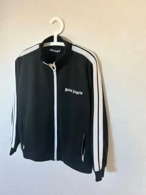 Svart ziptröja Palm Angels - svart ziptröja från Palm Angels med vita ränder längs ärmarna och logga på bröstet. Tröjan har hög krage, dragkedja framtill och fickor med dragkedja. Nästan aldrig använd, säljs då den är för liten för mig