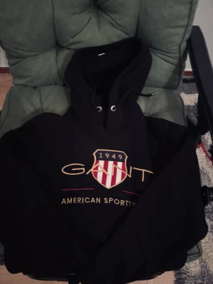 Svart hoodie från Gant med tryck - Svart hoodie från Gant med stort tryck på bröstet där det står 'GANT 1949 American Sportswear' och ett rött och vitt sköldmärke. Hoodien har snörning i huvan och ribbade muddar. Perfekt för en chill och sportig look.