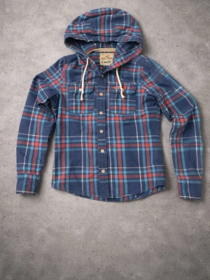 Blårutig hoodie från Hollister - Snygg blårutig skjorttröja från Hollister med huva och vita dragsnören. Tröjan har långa ärmar, två bröstfickor med knapp och klassiska knappar framtill. Perfekt för en avslappnad streetstil.