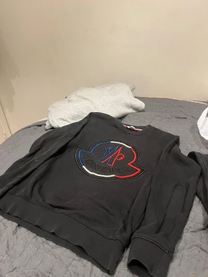 Svart sweatshirt från Moncler - Säljer en svart sweatshirt från Moncler med stort broderat logotypmärke i blått, rött och vitt på bröstet. Tröjan har rund halsringning och ribbade muddar vid ärmslut och nederkant. Perfekt för dig som gillar streetwear och exklusiva märken.