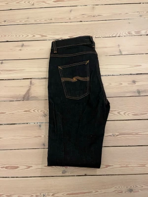 Mörkblå jeans från Nudie Jeans - Säljer ett par mörkblå jeans från Nudie Jeans med klassisk femficksmodell och kontrastsömmar. Jeansen har en slim passform och är tillverkade i kraftigt denimtyg av bomull. Snygg detalj med Nudie-loggan broderad på bakfickan.